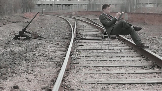 Un hombre con traje lee sentado en unas vías de tren